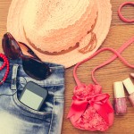 Summer women's accessories: sunglasses, beads, denim shorts, mobile phone, headphones, a sun hat, handbag, lipstick, nail polish. Toned image